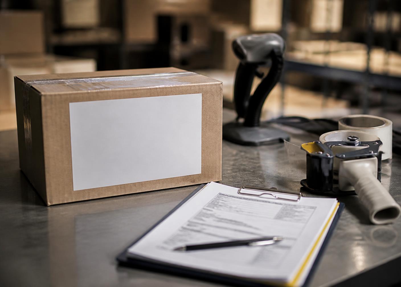 A packing station with a sealed carton, blank shipping label, packing list on a clipboard, scanner and tape gun.
