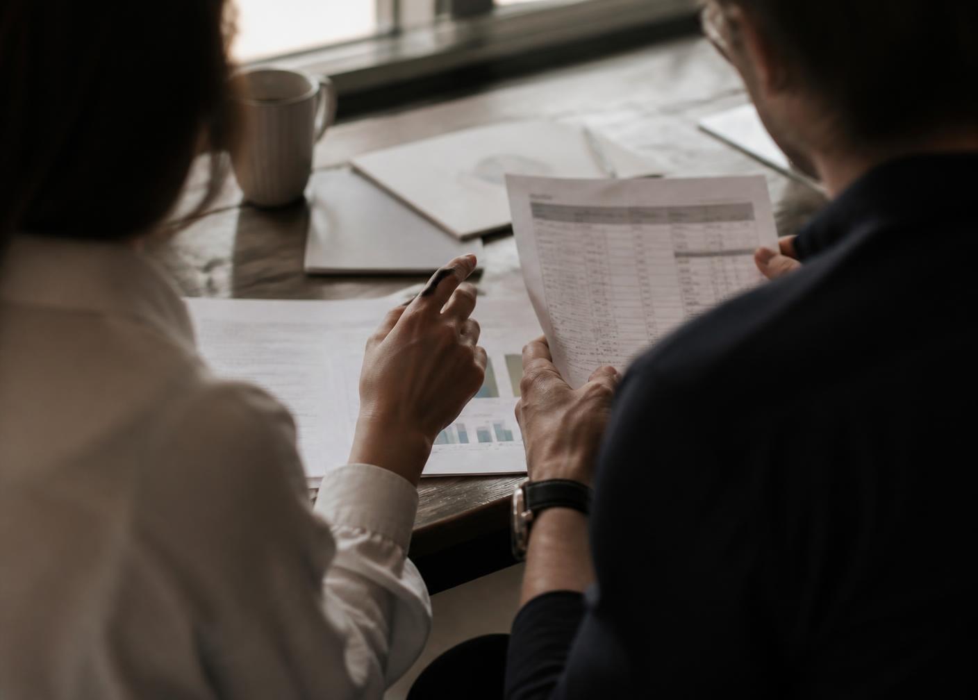 Two people seated at a table, seen from behind, reviewing printed pricing documents together.